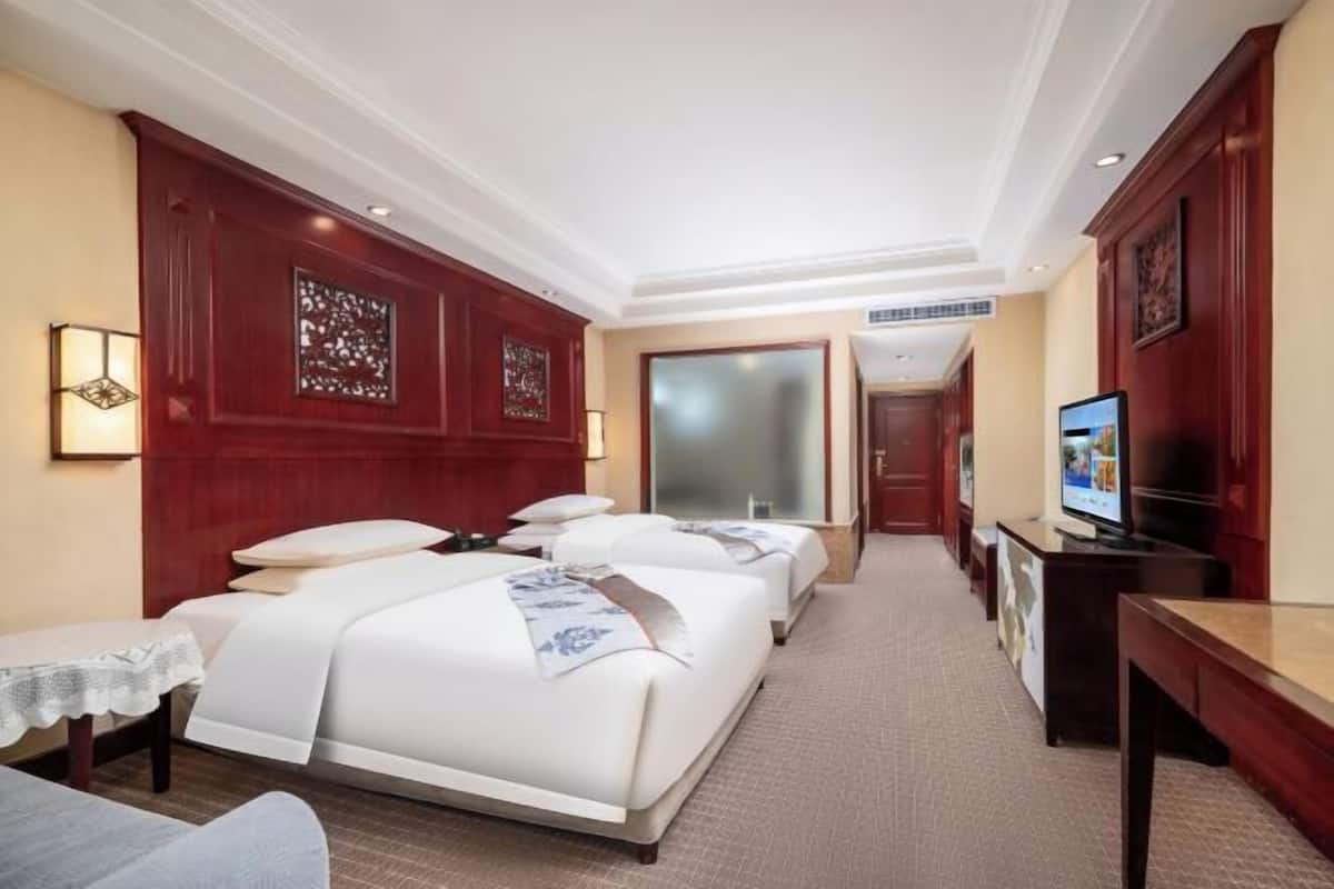 A hotel room with two beds, a desk, a TV, and a small table with a lamp.