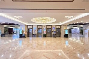 Un vestíbulo de hotel con piso de mármol, una araña central y varios ascensores.