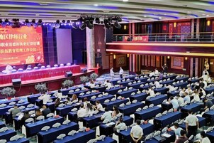 Un salón de conferencias con filas de mesas y sillas, un escenario con una pantalla grande y una zona de balcón.