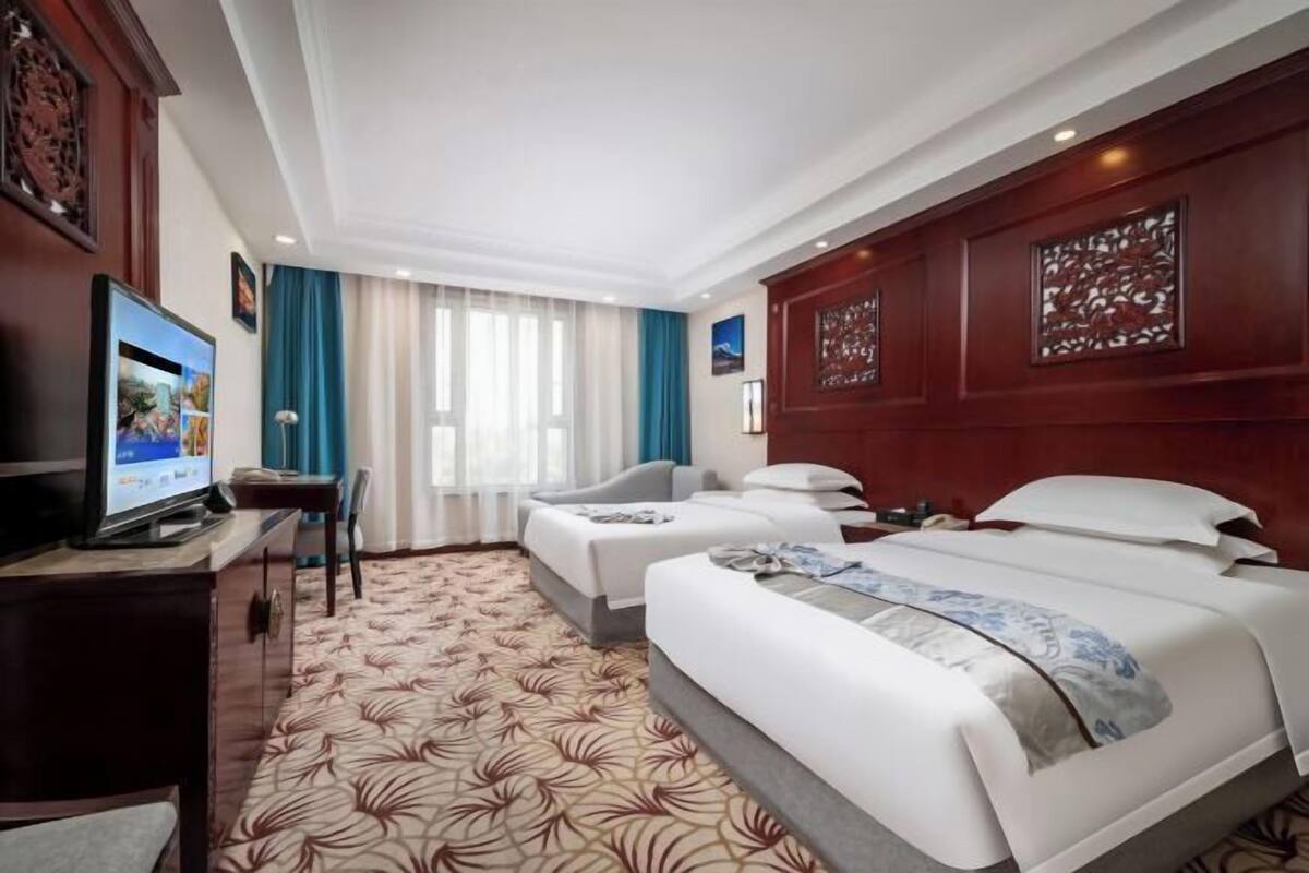 A hotel room with two beds, a desk, a TV, and a large window with curtains.