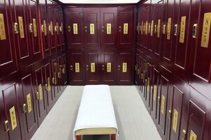 Una fila de taquillas de madera oscura con tiradores dorados y números, un banco blanco en el centro y un suelo alfombrado.