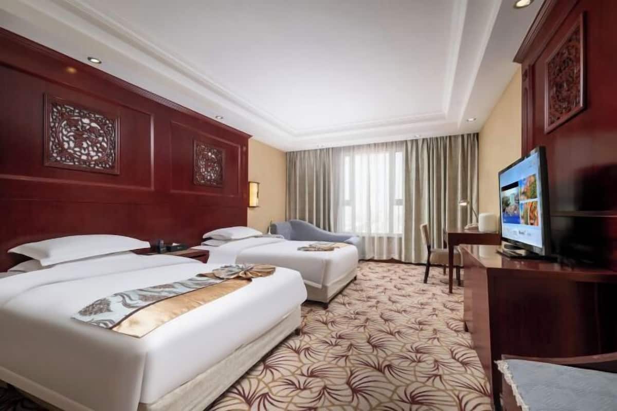 A hotel room with two beds, a TV, a desk, and a large window with curtains.