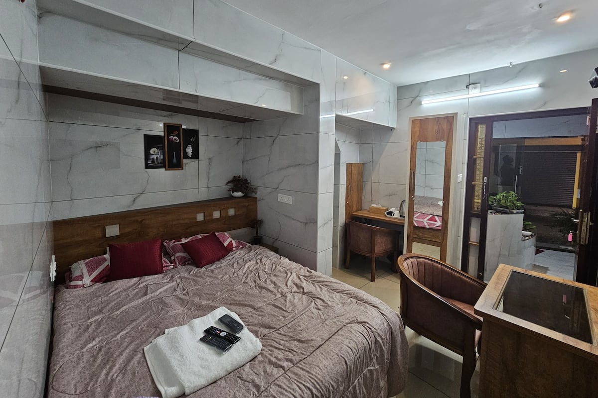 Un dormitorio moderno con pared de mármol, cabecero de madera, cama con ropa de cama roja y blanca, una mesita de noche, una silla y un pequeño escritorio con un espejo.