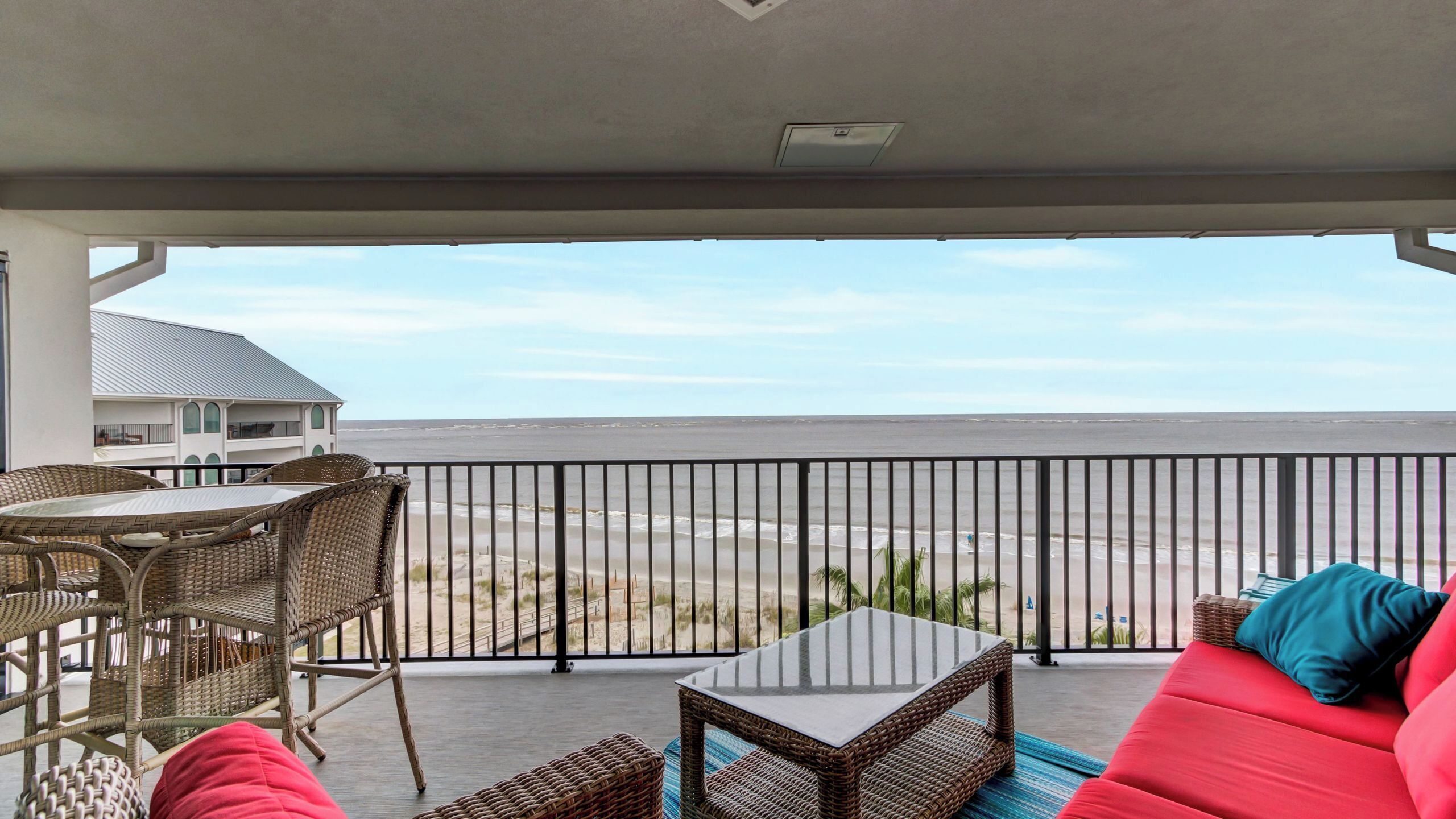 A balcony with wicker furniture, a glass table, and a red sofa with a view of the beach and ocean.
