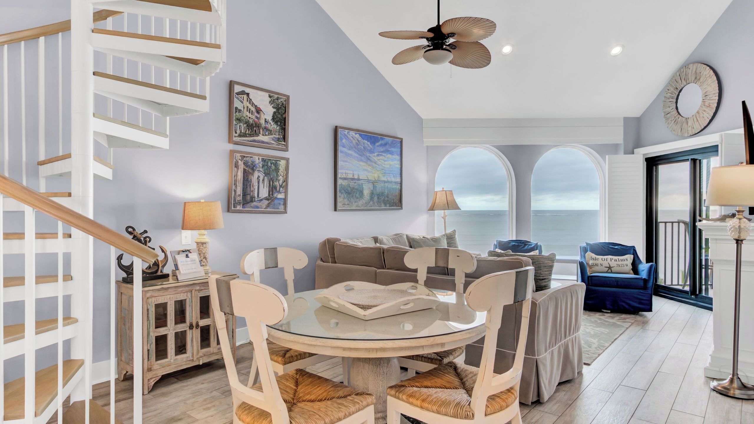 A spacious living area with a round glass table, a sofa, and a dining table with chairs. There is a staircase with a wooden handrail and a ceiling fan.