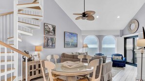 A spacious living area with a round glass table, white chairs, a sofa, and a staircase. There are framed pictures on the walls, a ceiling fan, and a view through large windows. - 518 Seascape Waterfront Views + Community Pool (Isle of Palms)