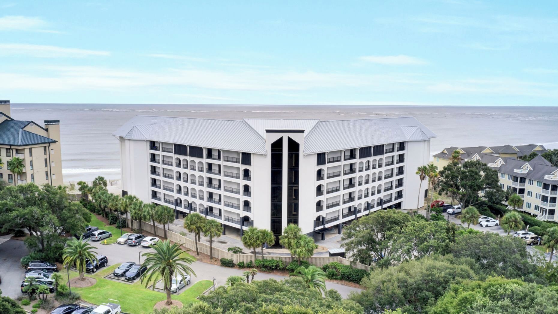 Een hotel met meerdere verdiepingen, een centraal atrium, een parkeerplaats en uitzicht op het strand.