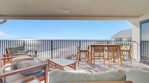 A balcony with a view of the beach, wooden furniture, and a glass table.