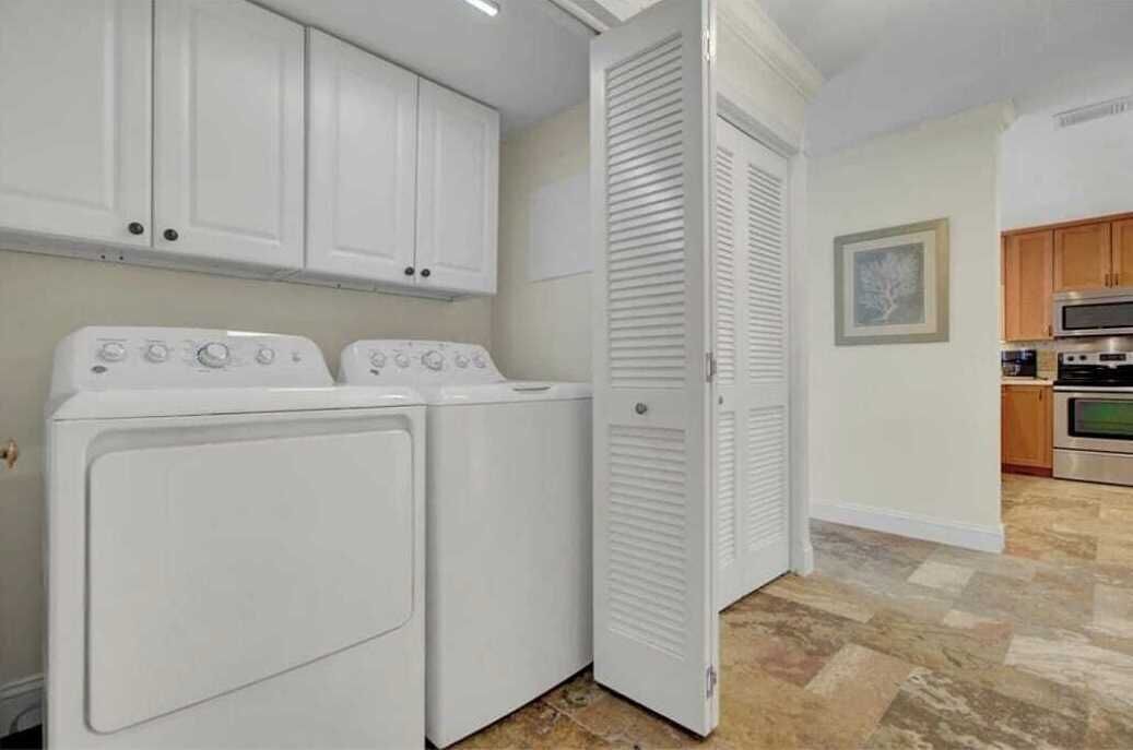 A laundry room with two white washers and dryers, a closed closet, and a kitchen area with wooden cabinets and stainless steel appliances.