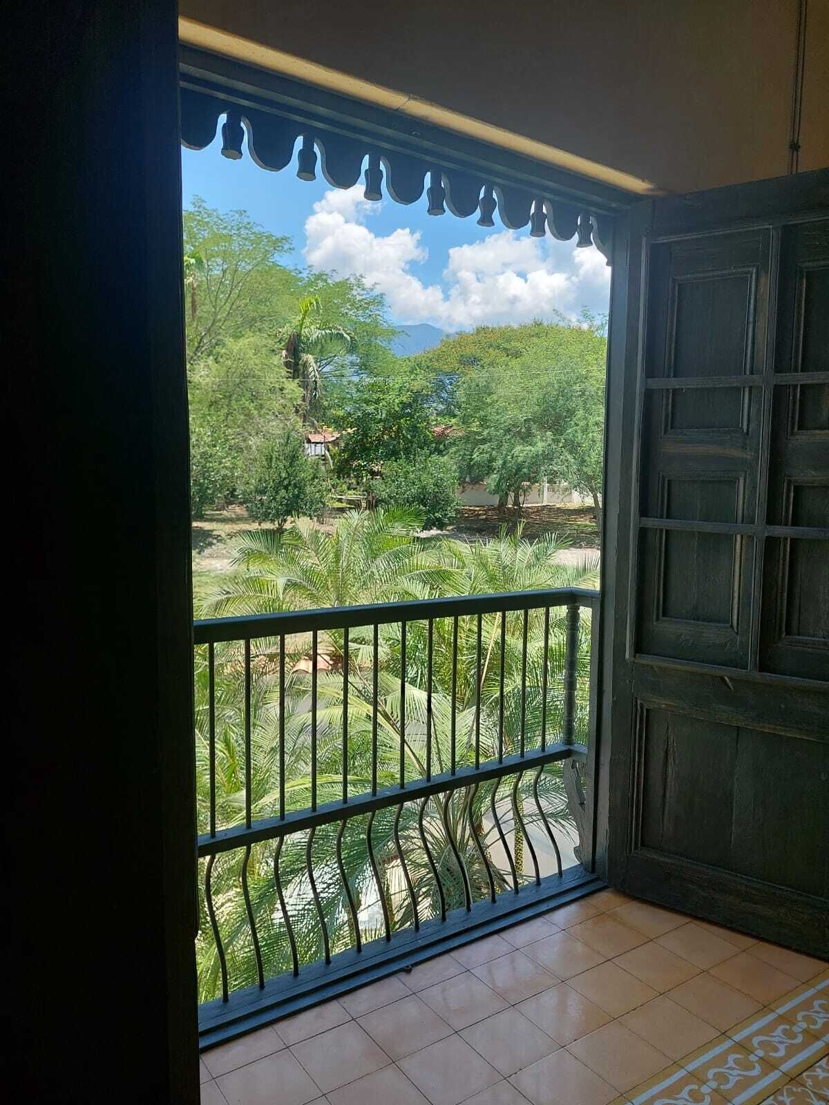 A room with a balcony featuring a metal railing, a tiled floor, and a view of a green landscape with trees and a clear sky.