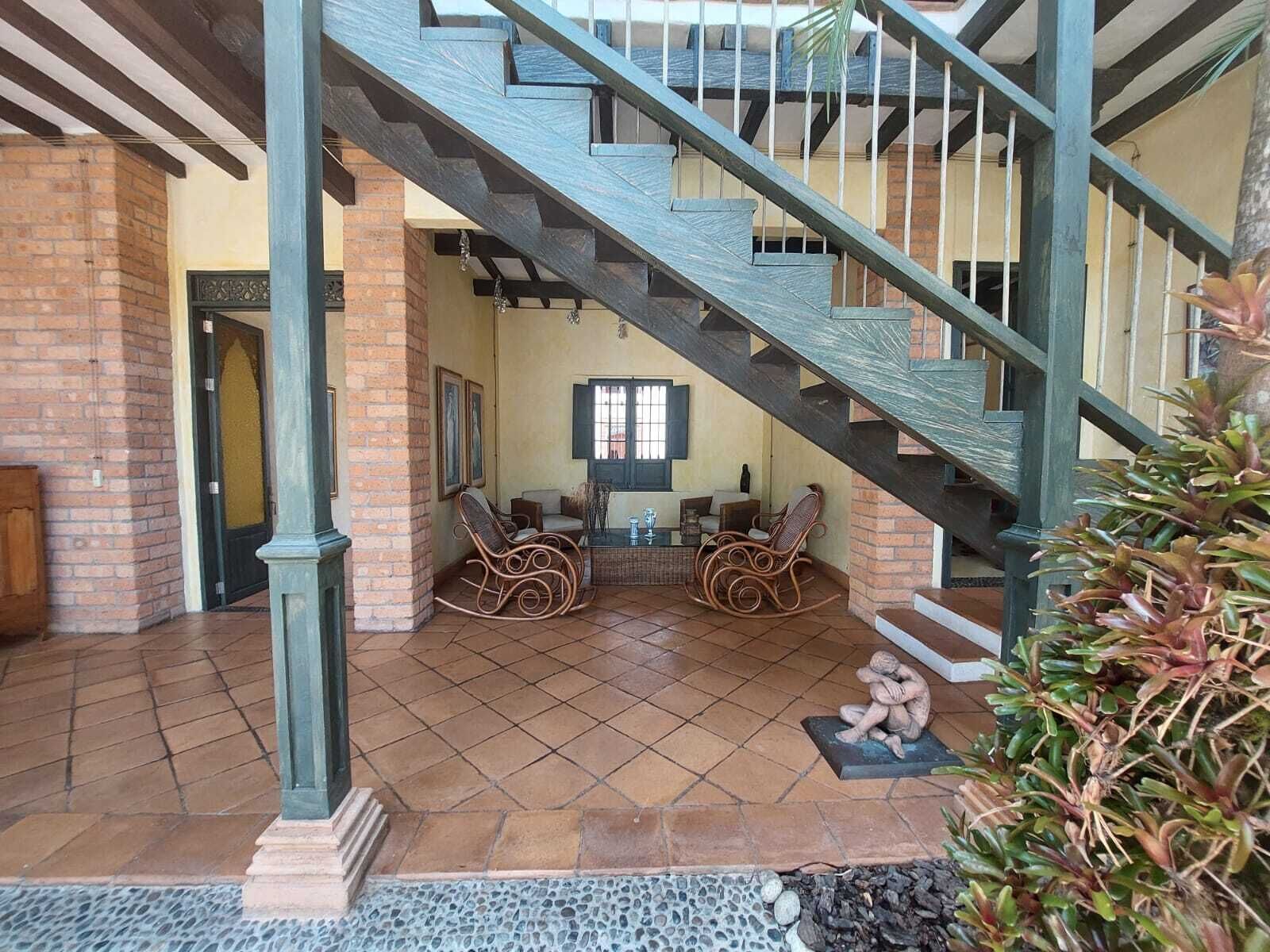 Un amplio espacio interior con una escalera de madera, una zona de estar con sillas de mimbre y un suelo de baldosas.