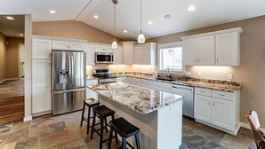Une cuisine moderne avec des meubles blancs, des appareils en acier inoxydable, un plan de travail en granit et des luminaires suspendus.