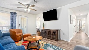A modern living room with a ceiling fan, a flat-screen TV mounted on the wall, a wooden coffee table, and a colorful rug.