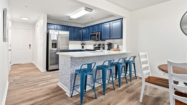 A modern kitchen with blue cabinets, a stainless steel refrigerator, a microwave, and a bar area with blue bar stools.