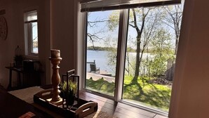 A room with a large sliding glass door leading to a deck overlooking a lake, wooden flooring, and a small table with a vase and plants.