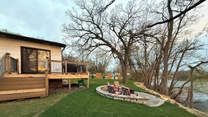 A deck with a view of a lake, a grassy lawn, and a patio with chairs.