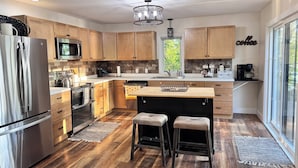 A modern kitchen with wooden cabinets, stainless steel appliances, a large island with stools, and a view through the window.