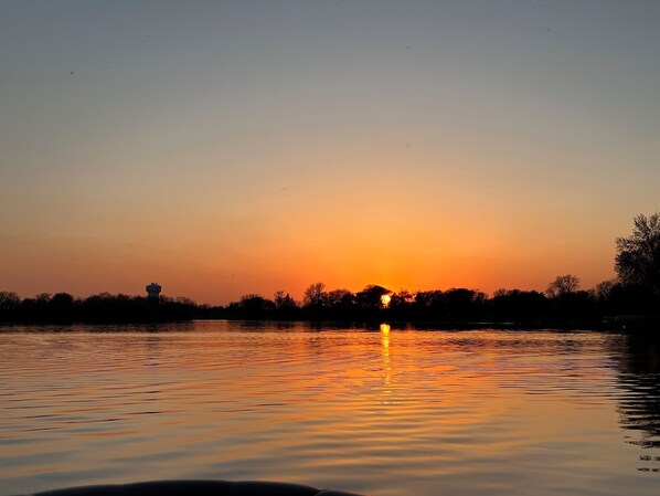 Atardecer sobre aguas tranquilas, con silueta de árboles y una estructura distante.