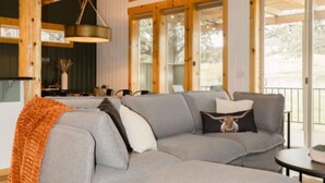 A modern living room with a grey sectional sofa, wooden beams, large windows, and a wooden floor. - Luxury Chalet + Arcade  3BRD Dogs OK (Redmond)