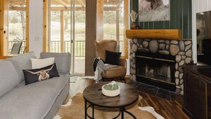 A cozy living room with a stone fireplace, a grey sofa, a leather armchair, and a large window overlooking a grassy area. - Luxury Chalet + Arcade  3BRD Dogs OK (Redmond)