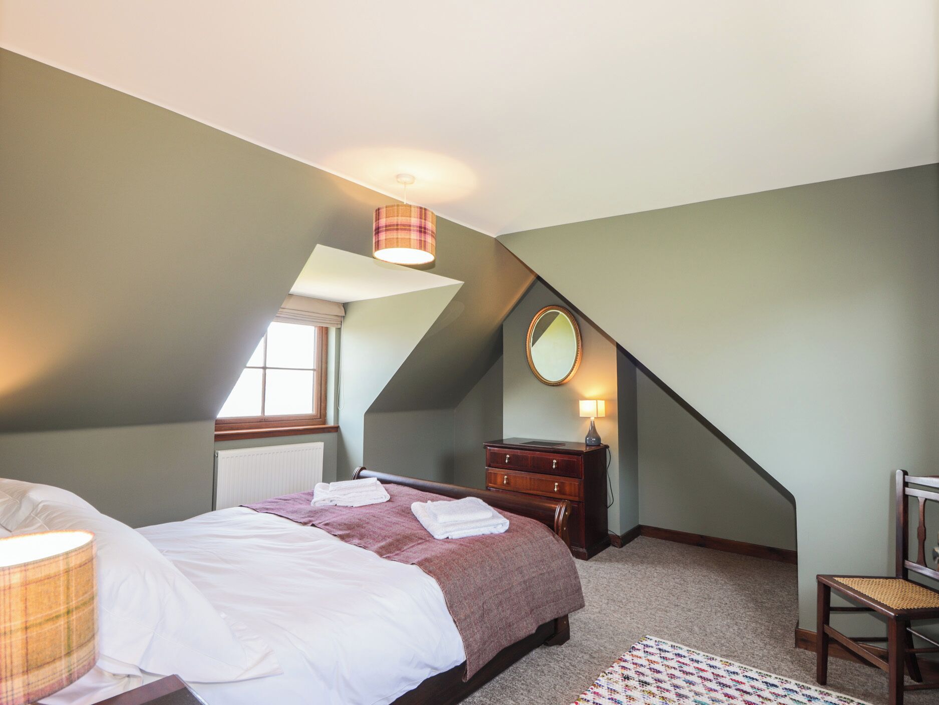 Ein Schlafzimmer mit schrägem Dach, einem Bett mit weißen Leinen, einer hölzernen Kommode, einem Spiegel und einem Fenster mit Jalousien.
