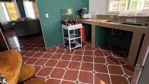 A compact kitchen with a tiled floor, a dining area with a wooden table and chairs, and a window with curtains.