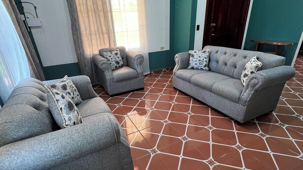 A living room with two grey sofas, patterned cushions, a window with curtains, and a tiled floor.