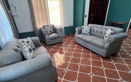A living room with two grey sofas, patterned cushions, a window with curtains, and a tiled floor.