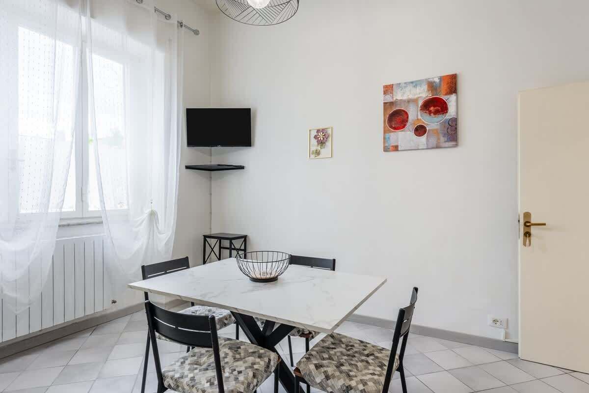 Un comedor con una mesa blanca, sillas negras, un televisor en la pared y obras de arte en la pared.