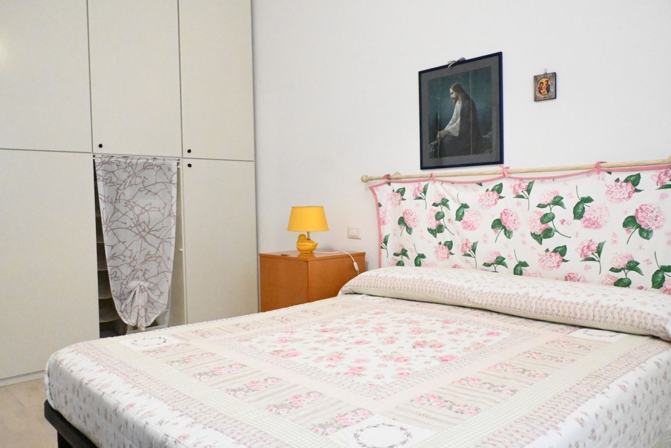 A bedroom with a bed, a nightstand with a lamp, a wardrobe, a framed picture on the wall, and a floral-patterned headboard.