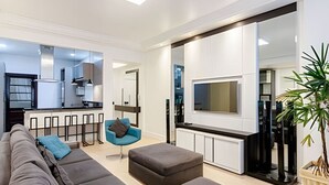 A modern living room with a sofa, ottoman, and a blue chair. A TV is mounted on the wall, and there is a kitchen area visible in the background. - Apartamento 3 Quartos 116m² no Batel Curitiba (Curitiba)