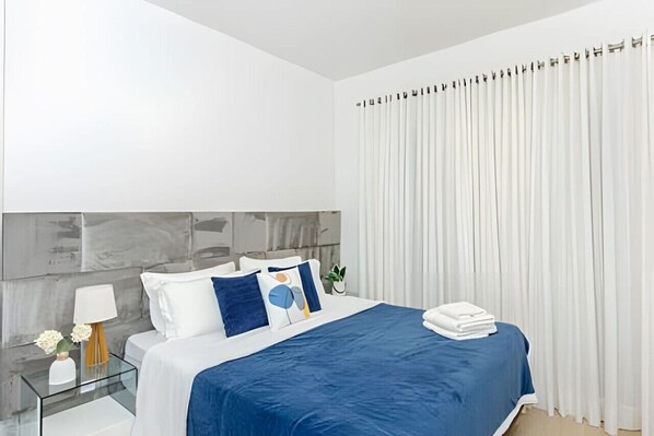 A modern hotel room with a large bed, a glass side table, a nightstand, and a white curtain.