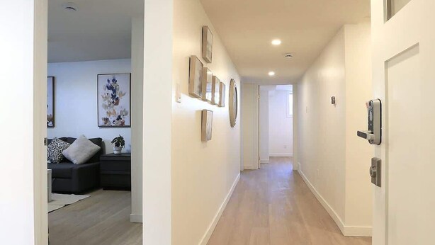 Een hal met houten vloer, witte muren en in het plafond weggewerkte verlichting. Er hangen ingelijste foto's aan de muren en aan de rechterkant is een deur met een modern slot.