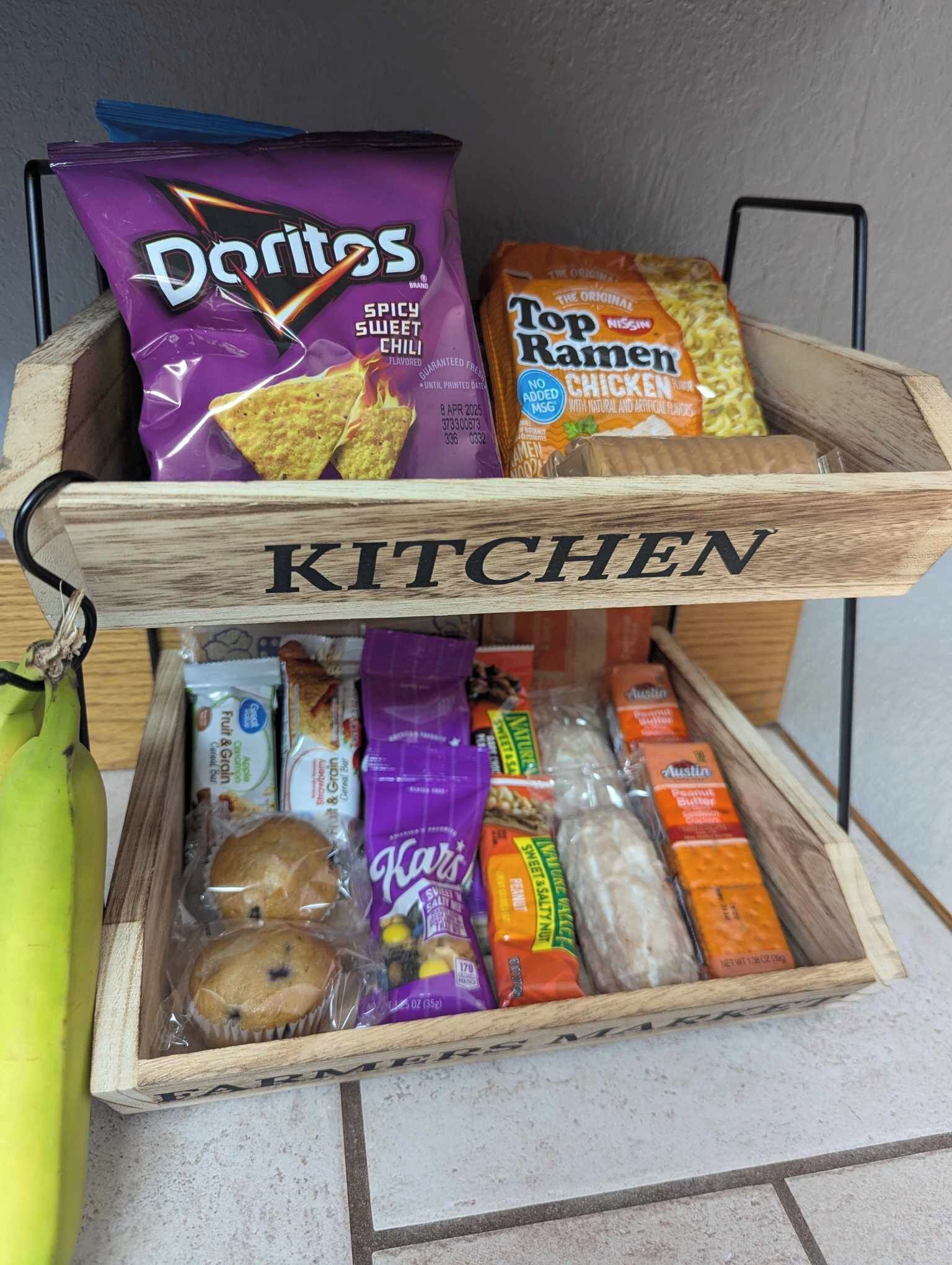 A wooden kitchen storage box labeled "KITCHEN" containing various food items like Doritos, Top Ramen, and snacks.