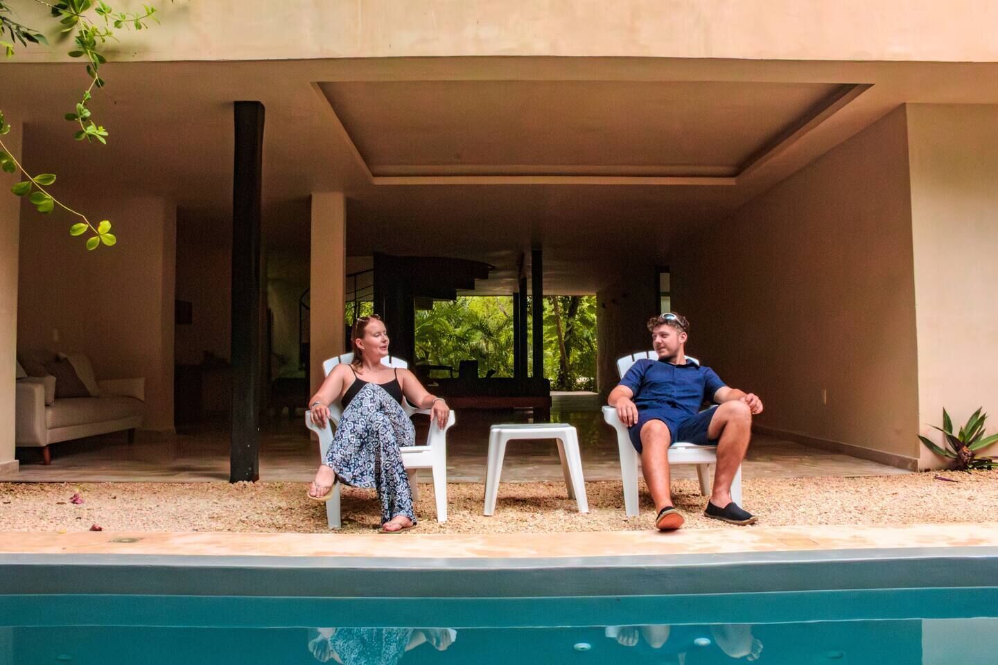 Two people sitting by a poolside in a covered outdoor area with a view of greenery in the background.