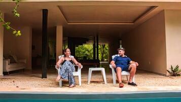 Two people sitting by a poolside in a covered outdoor area with a view of greenery in the background.