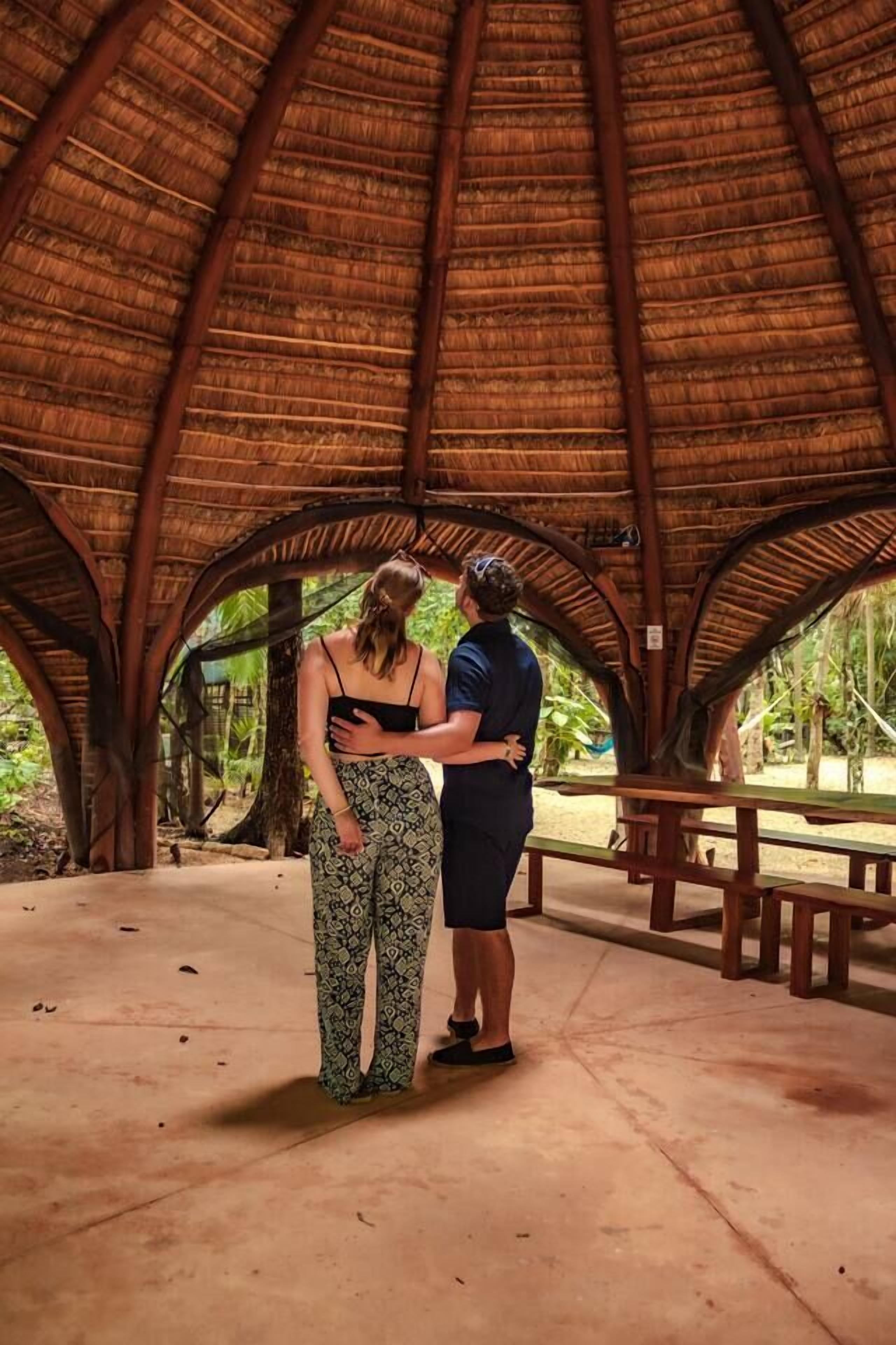 A covered outdoor area with a thatched roof, benches, and a concrete floor.
