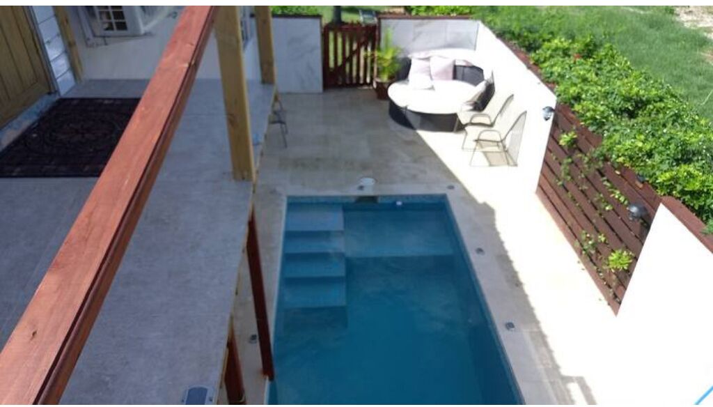 A small outdoor swimming pool with steps leading into it, surrounded by a wooden deck and a seating area with a table and chairs.