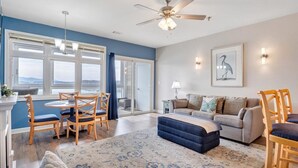 A spacious living room with a large window, a ceiling fan, a sofa, and a dining area with chairs. - #344 Pointe - 2 Bedroom Lakefront Condo at Mariners SML (Bedford County)