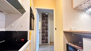 Een smalle keuken met een magnetron, oven en een kleine koelkast. Achtergrond is een boekenplank zichtbaar.