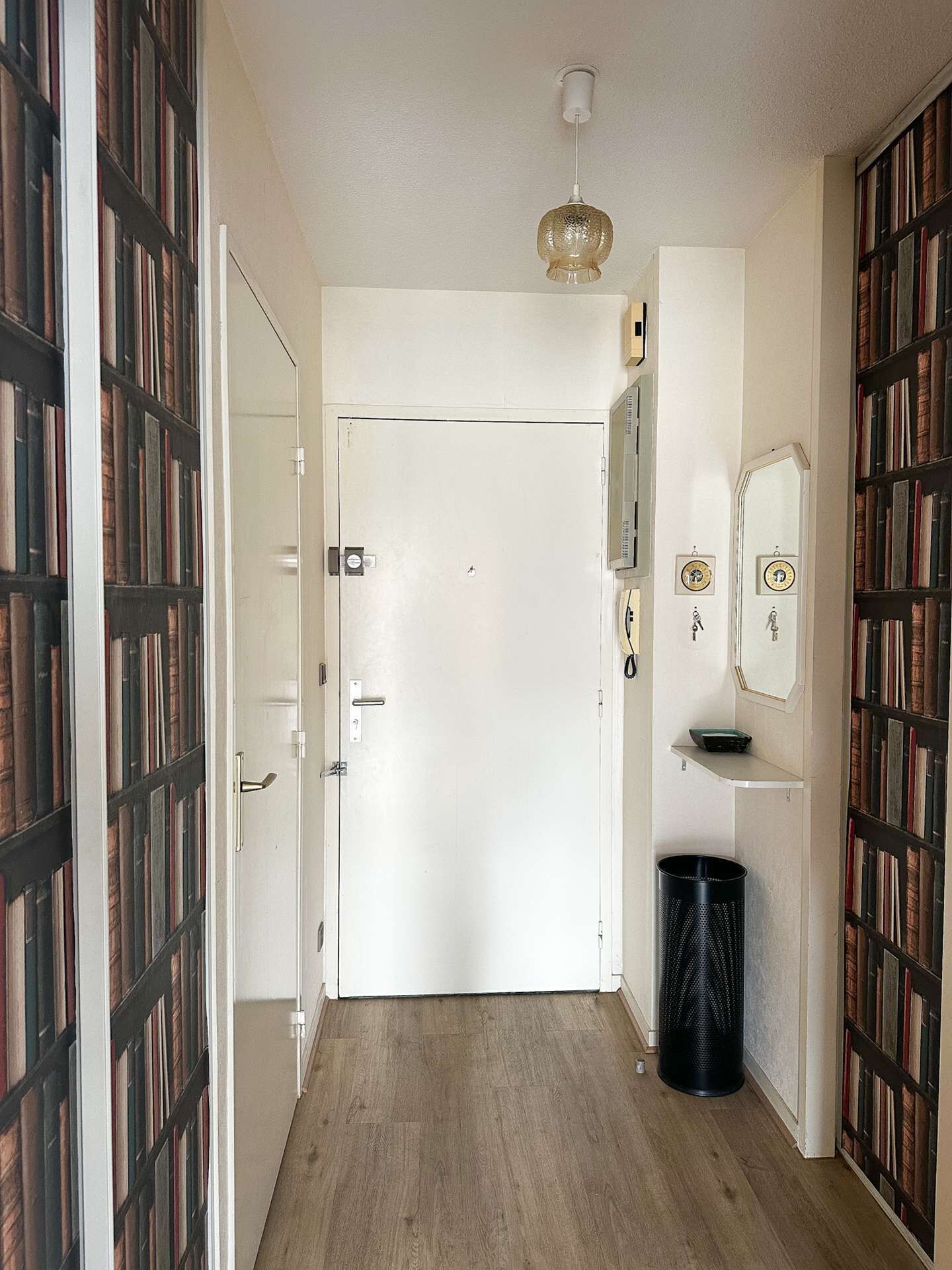 A hallway with a white door, wooden flooring, a black trash bin, and bookshelves on both sides.