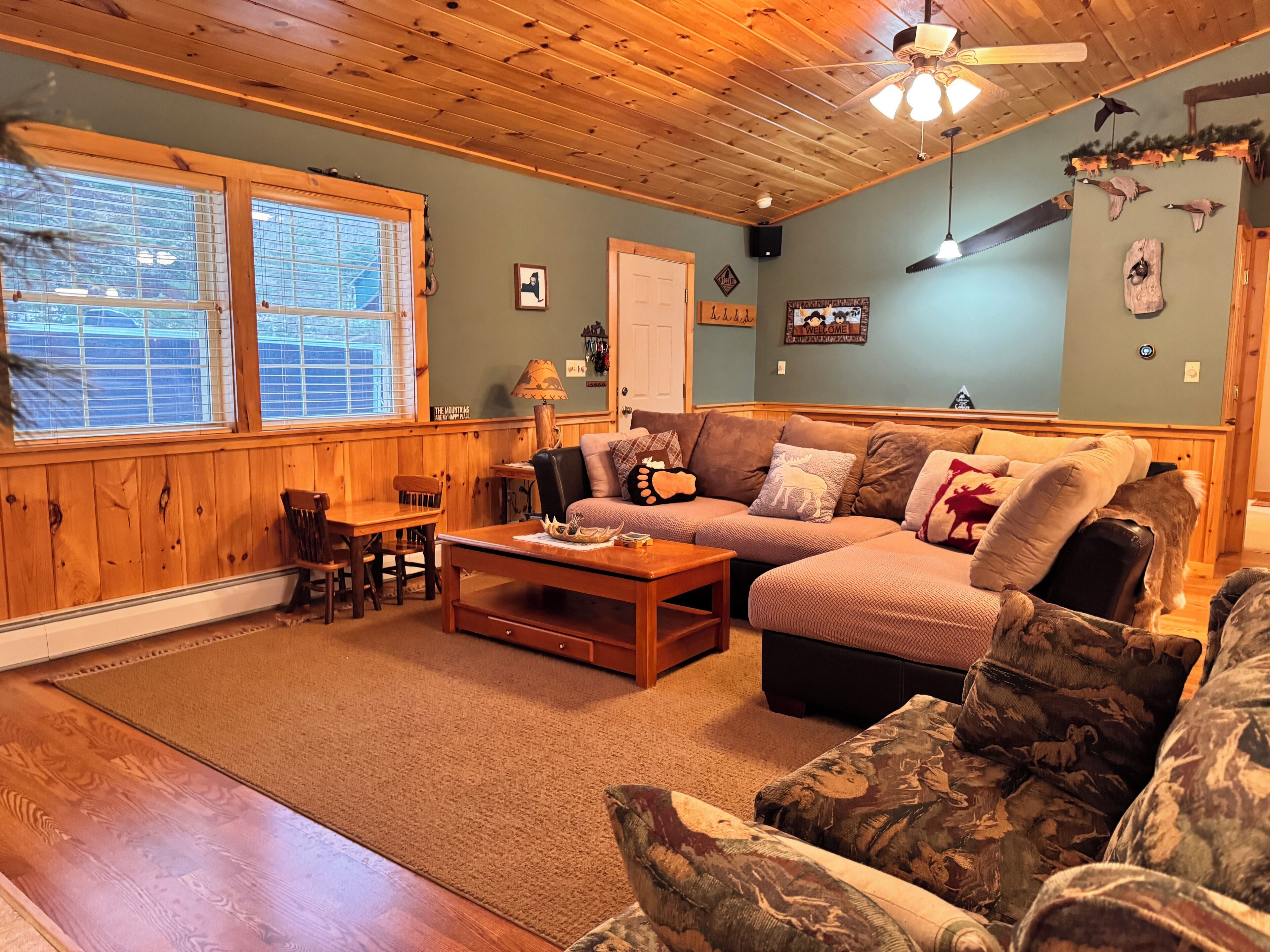 A cozy living room with wooden walls and ceiling, a brown leather sofa, a wooden coffee table, and a large window with blinds.