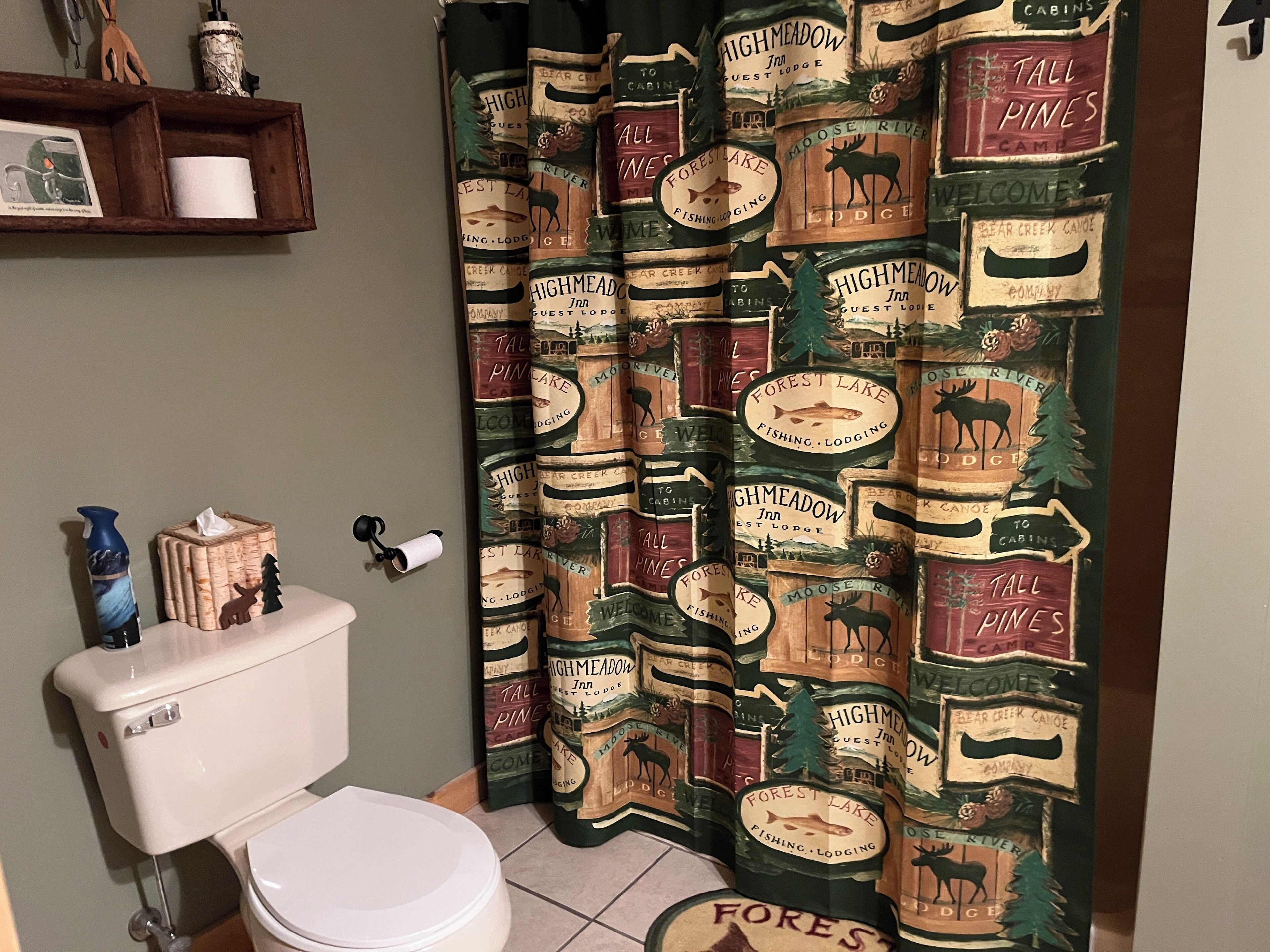 A bathroom with a toilet, a shelf with toiletries, and a shower curtain with forest and lodge designs.