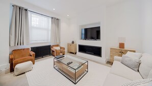 Ett modernt vardagsrum med en soffa, ett soffbord i glas, en TV som är monterad på väggen, en öppen spis och ett element.