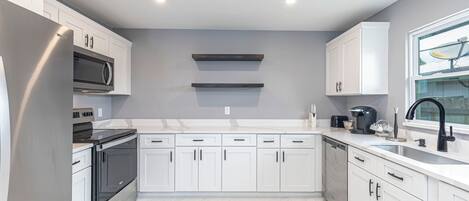 Eine moderne Küche mit weißen Schränken, einem Edelstahl-Kühlschrank, einem eingebauten Backofen, einer Mikrowelle und einem Geschirrspüler. Es gibt zwei Hängeschränke und ein Fenster mit Blick nach draußen.
