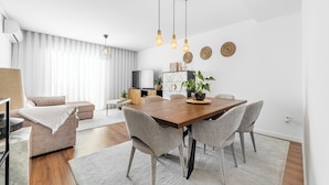 Een moderne woonkamer met een houten eettafel, grijze gestoffeerde stoelen en een groot raam met dunne gordijnen.
