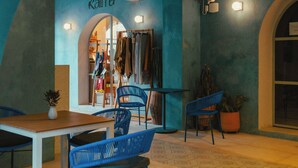 Een café met een blauwe boog, een houten tafel, blauwe stoelen en een kledingwinkel genaamd 'Kala' op de achtergrond.