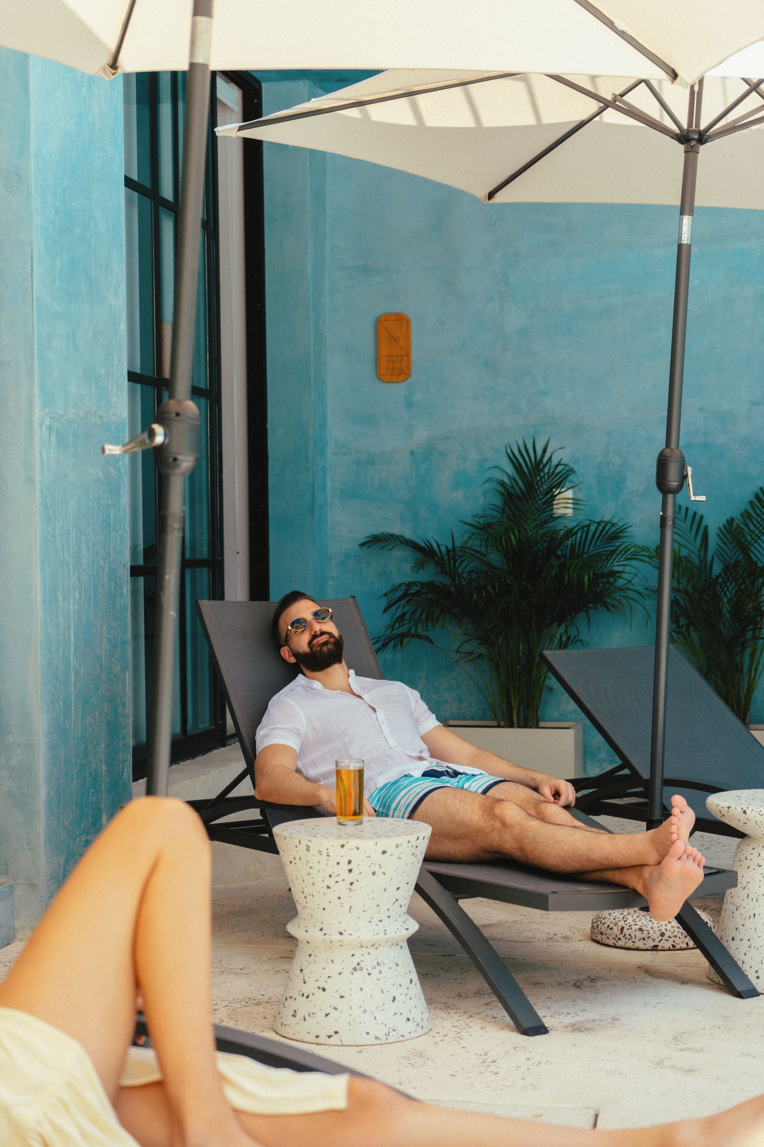 A man relaxing on a sun lounger with a drink, surrounded by a blue wall and potted plants.