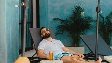 A man relaxing on a sun lounger with a drink, surrounded by a blue wall and potted plants.