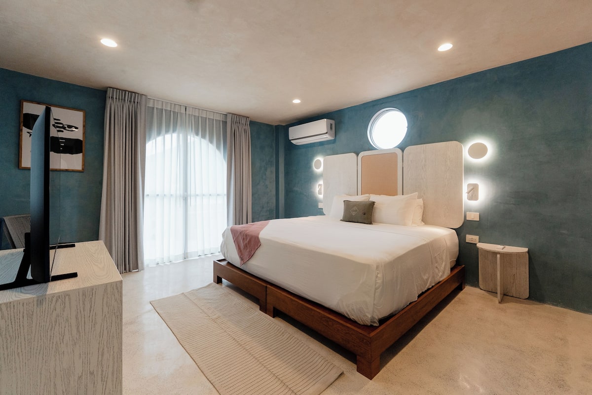 A modern bedroom with a large bed, a desk, and a vanity with lights.